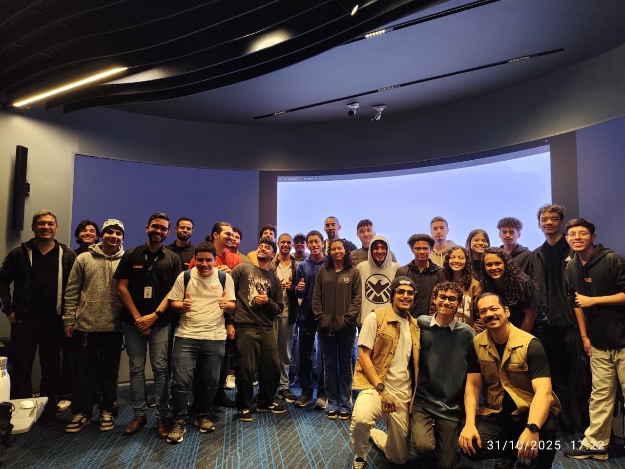 Os apresentadores e a turma da oficina reunidos diante da tela curva